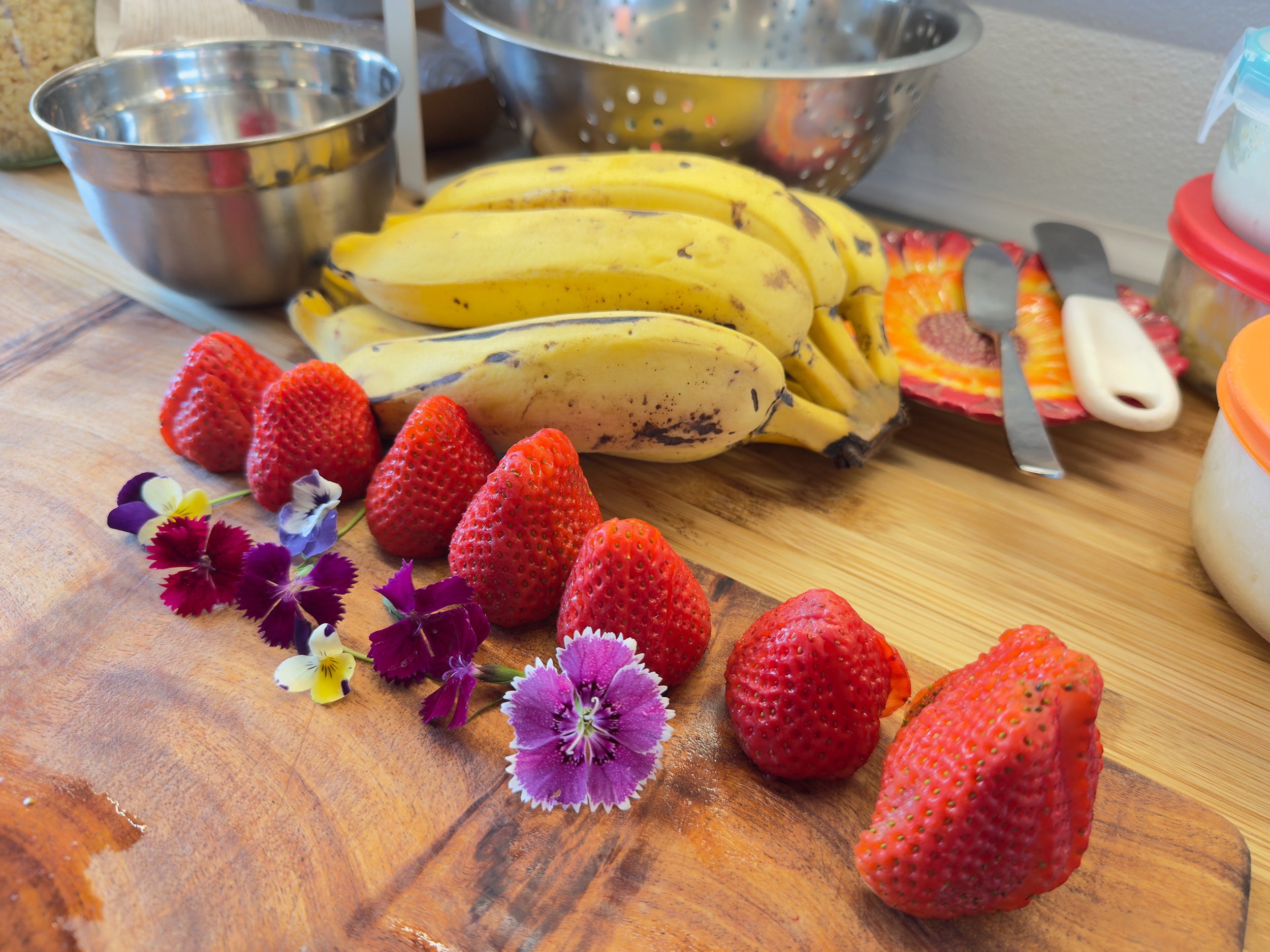 Aiwi Waffles toppings on a cutting board. A bushel of apple bananas and fresh strawberries sit on a cutting board alongside fresh flowers.
